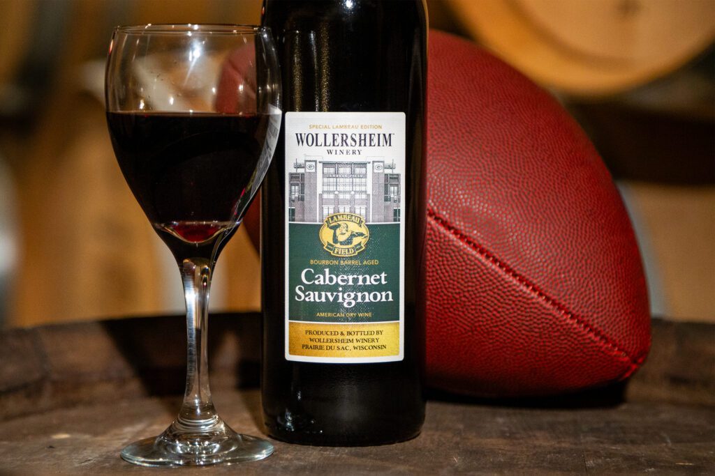 A bottle of Lambeau Edition Bourbon Barrel Aged Cabernet Sauvignon next to a glass of the wine and a football in the Wollersheim wine cellar surrounded by barrels of wine.