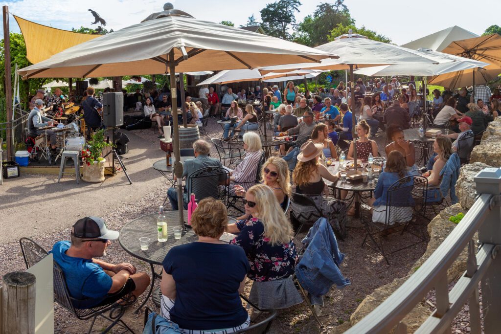 A large group of people enjoying drinks and food, listening to music on the Eagle Shelter patio at Wollersheim Summer Serenade.